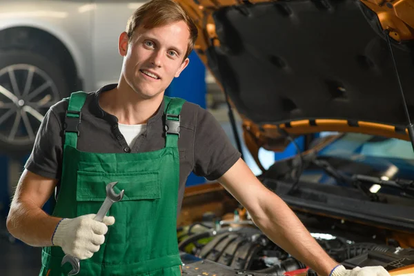 A young mechanic in green overalls holds a key in his hands near an orange car