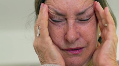 Yaşlı kadın gözlerini kapatıp şakaklarına masaj yapıyor, şiddetli baş ağrısı belirtileri gösteriyor. Onun acı ifadesine odaklan.