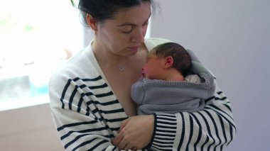 Anne, yumuşak bir battaniyeye sarılı olarak yeni doğan bebeğine bakıyor. Bebeği huzurlu ve sevgi dolu bir kucaklama içinde kucaklıyor. Derin bir bağ ve özen anının altını çiziyor..