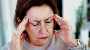 Olgun bir kadın şiddetli baş ağrısına katlanıyor, gözleri kapalı, elleri şakaklarda. İfade ve yüz hatları şiddetli acı, stres, zihinsel yorgunluk yükü, fiziksel veya duygusal gerginlik taşır.