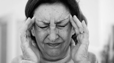 Görünür bir sıkıntı içindeki yaşlı kadın, iki eliyle yüzünü buruştururken, gözleri sımsıkı kapalı, yoğun bir şekilde ağrı kesiciye odaklanmış. Kırışıklıklar ve yüz ifadesi, baş ağrısı veya stres