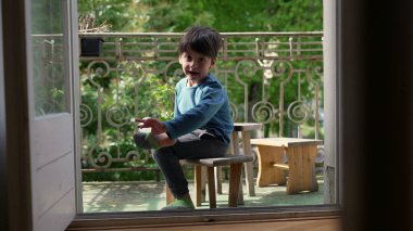 Genç çocuk bir balkonda tahta bir taburede oturuyor, gülümsüyor ve ayağını tutuyor, yeşilliklerle çevrili, yumuşak doğal ışıkla.
