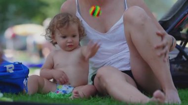 Bebek çimenlere atmadan önce renkli bir oyuncak tutuyordu ve yaz pikniğinde annesinin yanında duruyordu. Oyun dolu, açık havada geçen çocukluk anı merak ve enerjiyle doluydu.