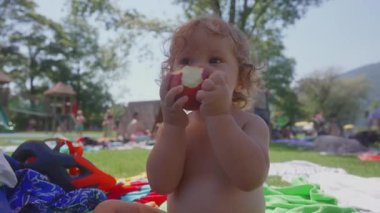 Yaz pikniği sırasında yeşil bir battaniyeye otururken ısırılmış şeftali tutan küçük çocuğun yakın çekimi, meyve yerken odaklanmış ifade, açık havada geçen samimi çocukluk anı