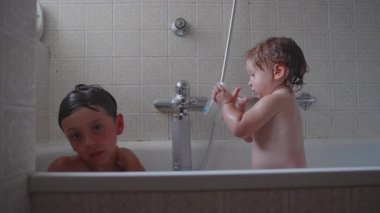 Ağabey ve yeni yürümeye başlayan çocuk küvette birlikte otururken, kucağında plastik bardakla kardeşlerin evde banyo yaparken neşeli, tatlı bir aile bağı kurdukları an.