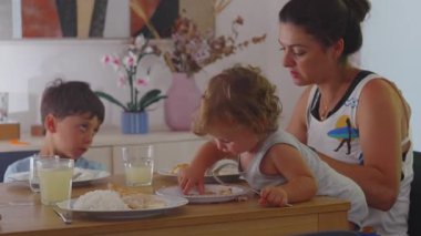 Anne evde iki çocukla öğle yemeği yiyor, anne yardım ederken kucağında kaşıkla öne doğru eğiliyor, aile yemeği zamanı, yanında kardeş ve masada yemek tabakları var.