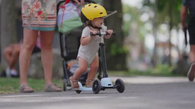 Sarı kasklı bir çocuk park yolunda mavi scooter sürüyor, gidonu sıkıca tutuyor ve meraklı bir şekilde yan bakıyor, çocukluk gezisi anı.