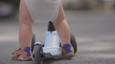 Mavi scooter 'ın üzerinde duran, sandalet giyen, denge, hareket ve çocukluk aktivitelerinin küçük detaylarını gösteren bebek bacaklarının ve ayaklarının yakın çekimi..