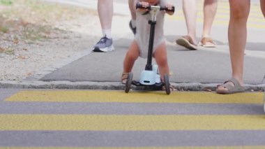 Sarı yaya geçidinde küçük mavi scooter kullanan bir çocuğun yakın çekimi, yayalar yakınlarda yürürken, hareketli bacaklar, hareketli ve hareketli şehir çocukluğu anı