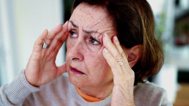 Olgun bir kadın şiddetli baş ağrısına katlanıyor, gözleri kapalı, elleri şakaklarda. İfade ve yüz hatları şiddetli acı, stres, zihinsel yorgunluk yükü, fiziksel veya duygusal gerginlik taşır.