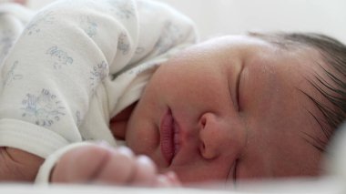 Serene uyuyan bebek, beyaz tulum içinde, bebeğin yüzünün ve elinin yakın çekimi, erken çocukluğun yumuşaklığını ve saflığını yakalıyor..