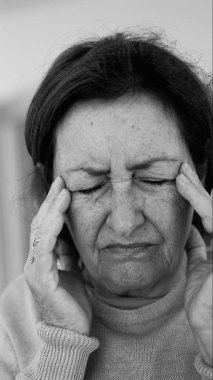 Yaşlı kadın gözlerini kapatıp şakaklarına masaj yapıyor, şiddetli baş ağrısı belirtileri gösteriyor. Onun acı ifadesine odaklan.