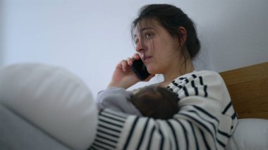Anne, yeni bebeği kollarında uyurken telefonda konuşuyor. Yorgunluk ve bağlılık karışımını iletiyor. Erken ebeveynlikte dinlenme ve sorumluluğun dengelendiği sakin anlar..