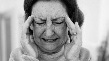 Görünür bir sıkıntı içindeki yaşlı kadın, iki eliyle yüzünü buruştururken, gözleri sımsıkı kapalı, yoğun bir şekilde ağrı kesiciye odaklanmış. Kırışıklıklar ve yüz ifadesi, baş ağrısı veya stres.