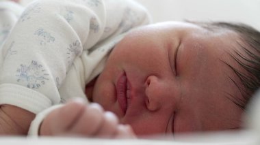 Serene uyuyan bebek, beyaz tulum içinde, bebeğin yüzünün ve elinin yakın çekimi, erken çocukluğun yumuşaklığını ve saflığını yakalıyor..