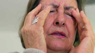 Kıdemli kadın gözlerini sıkıca kapatıyor ve iki eliyle tapınaklara masaj yapıyor. Acıyı ya da stresi azaltmaya odaklanıyor. Kırışıklıklar ve ifade, duygusal yükü vurgular.