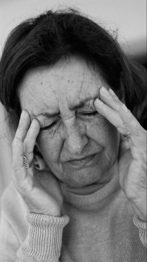 Yaşlı kadın gözlerini kapatıp şakaklarına masaj yapıyor, şiddetli baş ağrısı belirtileri gösteriyor. Onun acı ifadesine odaklan..