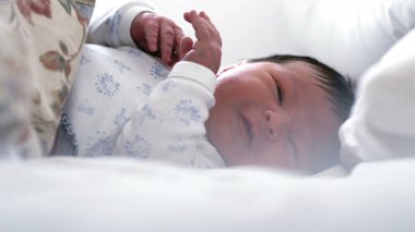 Yeni doğmuş bebek yatakta yatıyor, elleriyle jestler yapıyor ve yüzünü buruşturuyor. Bebek yumuşak yastıklarla çevrili, yatak odasında rahat ve huzurlu bir atmosfer yaratıyor..