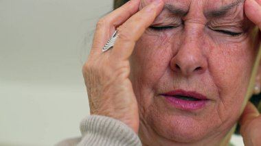 Kıdemli kadın gözlerini sıkıca kapatıyor ve iki eliyle tapınaklara masaj yapıyor. Acıyı ya da stresi azaltmaya odaklanıyor. Kırışıklıklar ve ifade, duygusal yükü vurgular.