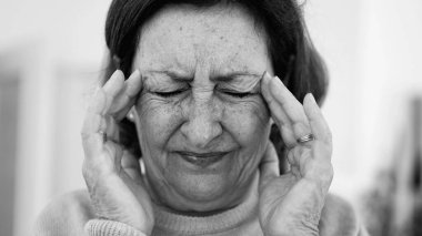 Görünür bir sıkıntı içindeki yaşlı kadın, iki eliyle yüzünü buruştururken, gözleri sımsıkı kapalı, yoğun bir şekilde ağrı kesiciye odaklanmış. Kırışıklıklar ve yüz ifadesi, baş ağrısı veya stres.