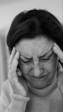 Yaşlı kadın gözlerini kapatıp şakaklarına masaj yapıyor, şiddetli baş ağrısı belirtileri gösteriyor. Onun acı ifadesine odaklan..