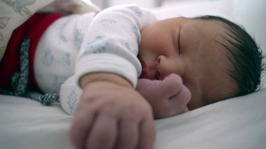 Yeni doğan bebek bir eli açık, diğer eli yüzü yakın bir şekilde bir yatakta huzur içinde uyuyor. Çocuğun sakin ifadesi ve rahat duruşu bir huzur ve güvenlik anını vurguluyor.