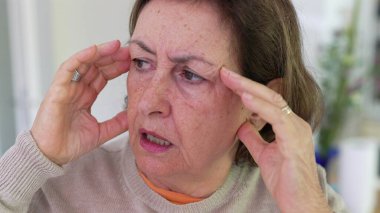 Büyük stres altındaki yaşlı kadın, parmaklarıyla tapınaklara masaj yapıyor, gözleri odaklı, yoğun endişe ve zihinsel yük altında. Detaylı ifade, duygusal gerginliğin etkisi veya fiziksel acı.