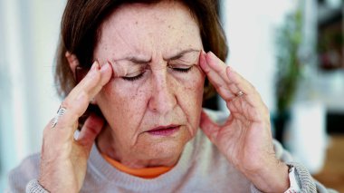 Olgun bir kadın şiddetli baş ağrısına katlanıyor, gözleri kapalı, elleri şakaklarda. İfade ve yüz hatları şiddetli acı, stres, zihinsel yorgunluk yükü, fiziksel veya duygusal gerginlik taşır..