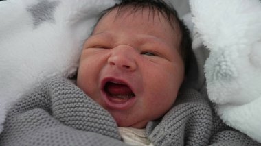 Yeni doğmuş bebek sıcak bir battaniyeye sarılmış, hafif kaşları çatık, rahatsızlık belirtileri gösteriyor, erken dönem duygularını yakalıyor, sıcak ve güvenli bir şekilde sarmalanıyor..