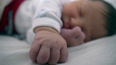 Yeni doğan bebek bir eli açık, diğer eli yüzü yakın bir şekilde bir yatakta huzur içinde uyuyor. Çocuğun sakin ifadesi ve rahat duruşu bir huzur ve güvenlik anını vurguluyor.