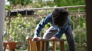 Genç Çocuk, balkonda tahta bir tabureye yaslanmış, iki elini de destek için kullanıyor, oyuna odaklanmış, elindeki göreve odaklanmış, doğal yeşillikle çevrili.