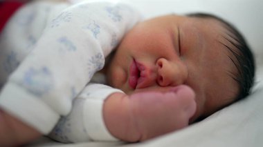 Yeni doğmuş bebek bir yatakta huzur içinde uyuyor. Nazik, rahat bir yüz ifadesi ve bir eli yüzünün yanında dinleniyor. Rahat bir ortamda huzurlu bir dinlenme ve rahatlık anı.