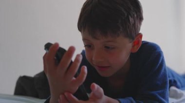 Akıllı telefonuyla yatakta uzanmış gülümseyen genç bir çocuk, ekranda rahatlamış ve mutlu bir çocukluk geçirirken keyifli bir anın tadını çıkarıyor.