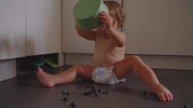 Gülümseyen çocuk mutfakta oturmuş yeşil kaseden yaban mersini düşürüyor, neşeli bir karmaşa anı, neşeli bir ifade, mutlu bir çocukluk ve ev ortamı