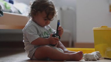 Kıvırcık saçlı çocuk yerde oturmuş oyuncak parçalarıyla oynuyor, odaklanmış ve meraklı, nesneleri keşfediyor ve evde oynayarak öğreniyor.