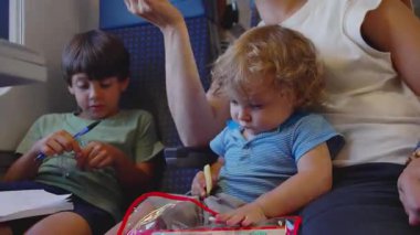 Küçük çocuk tren yolculuğu sırasında annesinin kucağında atıştırırken büyük kardeş de tren vagonunda rahat bir aile seyahati anı olan kalem ve plastik çantayla oynuyor.