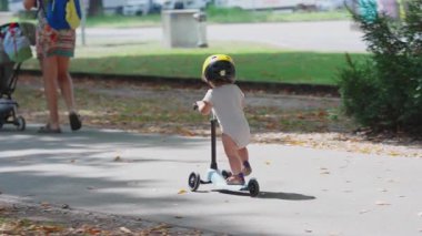 Güneşli park yolunda mobiletle gezen çocuk sarı kask takıyor, anne babayla bebek arabasının olduğu uzak bir manzara, eğlenceli ve tuhaf çocukluk aktivitesi