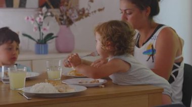 Küçük çocuk öğle yemeğini evde yerken, anne yakından izliyor, kardeş ile yemek masasında oturuyor, ev yapımı yemek sırasında aile bağlarını sıkılaştırıyor.