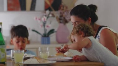 Anne, yeni yürümeye başlayan çocukla ve büyük kardeşiyle öğle yemeği yiyor. Bebek, kaşıkla öne doğru eğiliyor. Aile yemeği yiyor.