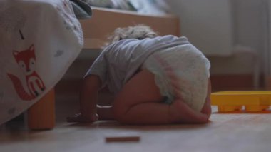 Kıvırcık saçlı çocuk oyuncak parçayı tutarken eğiliyor, yerde nesneleri keşfediyor, evde neşeli ve meraklı bir an.