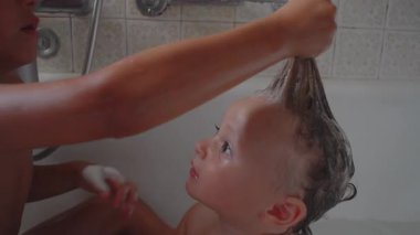 Büyük kardeş yıkanan bebeklerin saçlarını şampuan ve köpükle yıkıyor, çocuk güven ve merakla yukarı bakıyor, neşeli banyo zamanı aile bağlarını kuvvetlendiriyor..