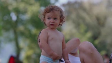 Yaz pikniği sırasında annesinin yanında çimlerin üzerinde duran meraklı çocuk, neşeli bir ifadeyle çevreyi gözlemliyor, masumiyetle dolu açık havada çocukluk anı..