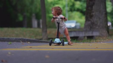 Küçük çocuk sarı yaya geçidinde yeşil ağaçlar ve yumuşak yaz atmosferiyle çevrili sakin bir banliyö caddesinde küçük mavi scooter sürüyor.