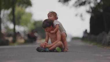 Yürüyen bebek, parkta yan yana otururken ağabeyine arkadan sarılıyor. Kaygısız yaz öğleden sonrasında kardeşler arasında sevgi dolu bir bağ kuruyor..