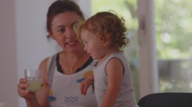 Anne, bebeğe evde meyve suyu içmesi için yardım ediyor, yemek masasında oturuyor, rahat ve samimi bir aile anı yaşıyor.