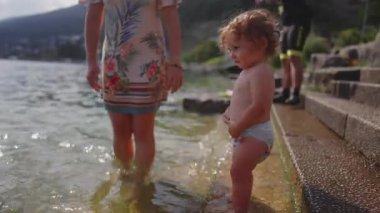 Sığ göl suyunda durup annelerin elini tutan, sonra taş basamakları tırmanmaya çalışırken, özgürlüğünü arayan, merakla çevreyi keşfeden çocuk.