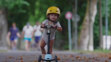 Sarı kask takan çocuk park yolunda scooter sürüyor. Gidonu sıkı sıkı tutuyor. Ağaçlarla çevrili ve arka planda bulanıklık var.