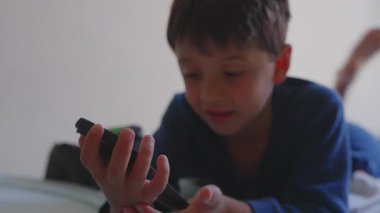 Mutlu genç çocuk, akıllı telefonu tutarak yatakta uzanıyor, ekranlara bakarken gülümsüyor, evde neşeli ve rahat bir çocukluk anı.