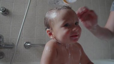 Anne, şampuanı temizlemek için konteynırdan su dökerek bebeklerin saçlarını duruluyor. Çocukla banyo yapıyor ve evde aile bağı kuruyor.