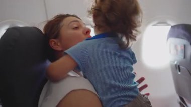 Anne gündüz uçuşu sırasında uçakta otururken huzursuz, kıvırcık saçlı bir çocuğu kucağına alıp zor bir aile seyahati sırasında telaşlı bir çocuğu teselli etmeye çalışıyor.